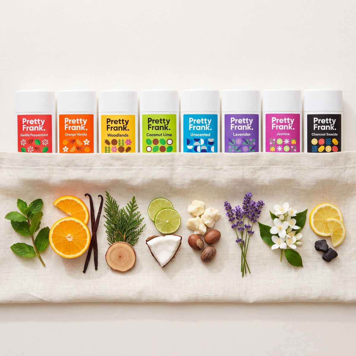 All eight Pretty Frank deodorant sticks lined up above the real ingredients that scent them: mint, orange, vanilla, cypress, lime, coconut, shea butter, lavender, jasmine, lemon and charcoal